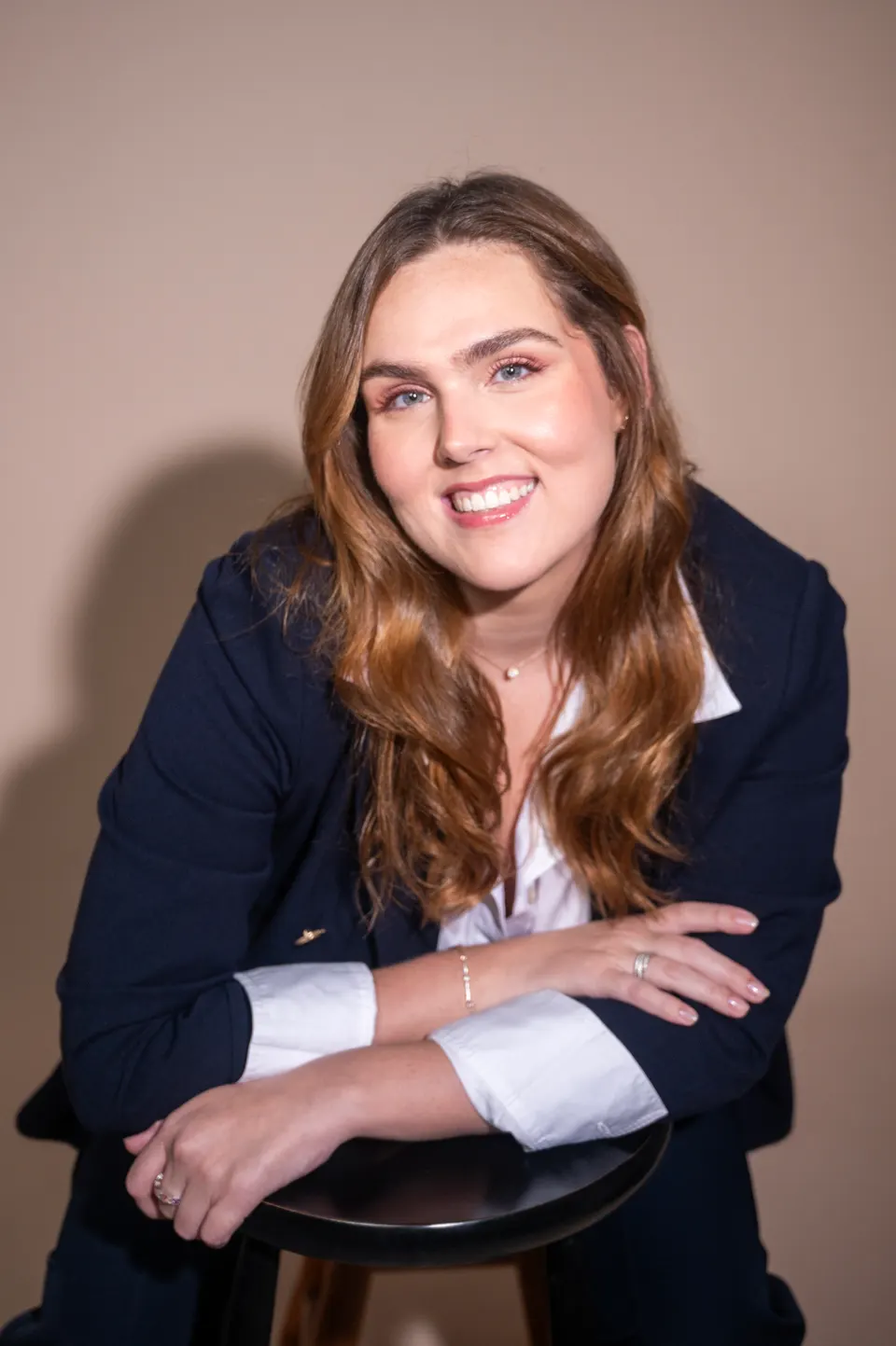 Mulher de cabelos longos e blazer escuro, sorrindo apoiada em um banco
